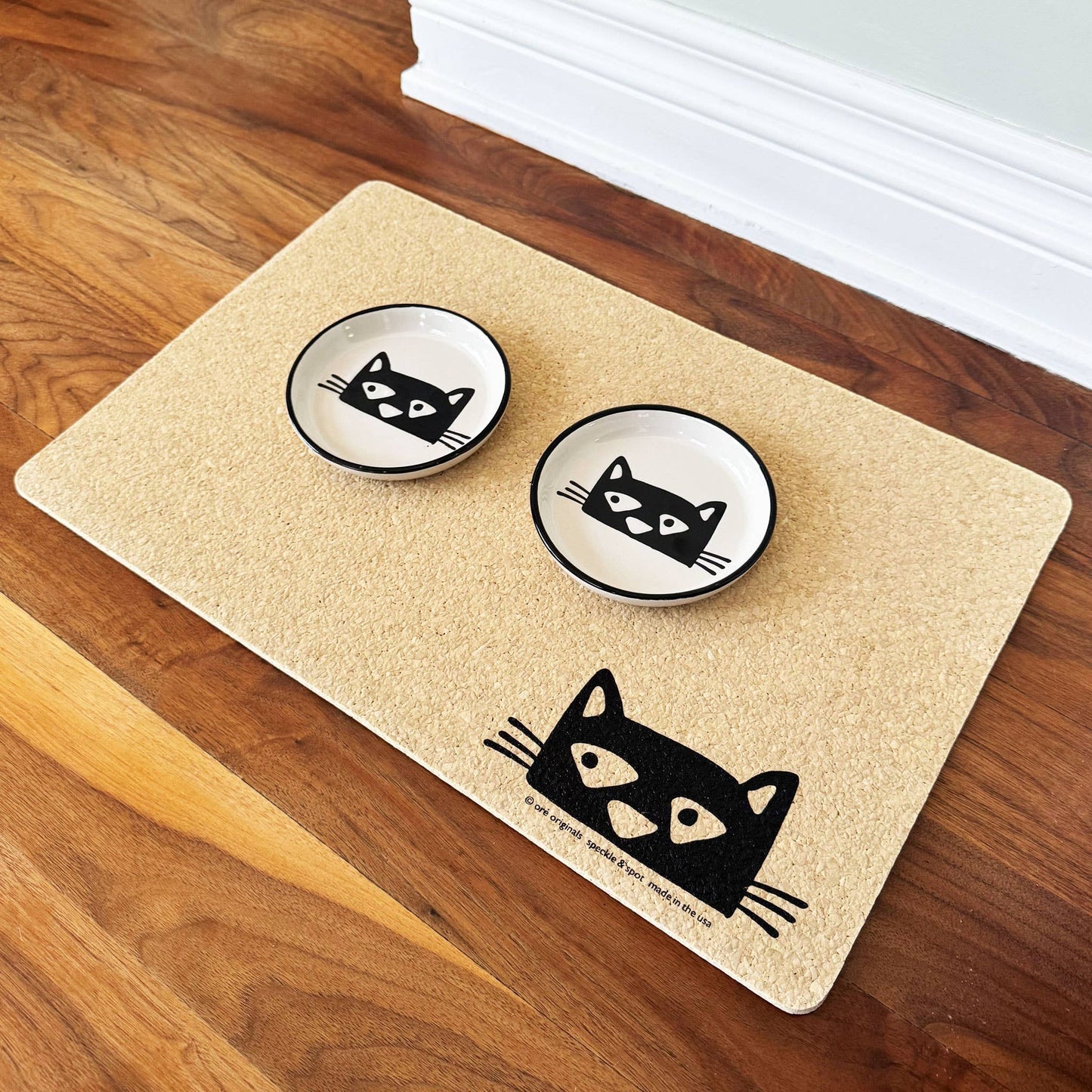 Pet mat with two bowls and a cat design on a wooden floor.