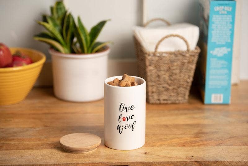 A white ceramic airtight treat jar with a bamboo lid, labeled 'live love woof', placed on a wooden surface, with a plant and a basket in the background.