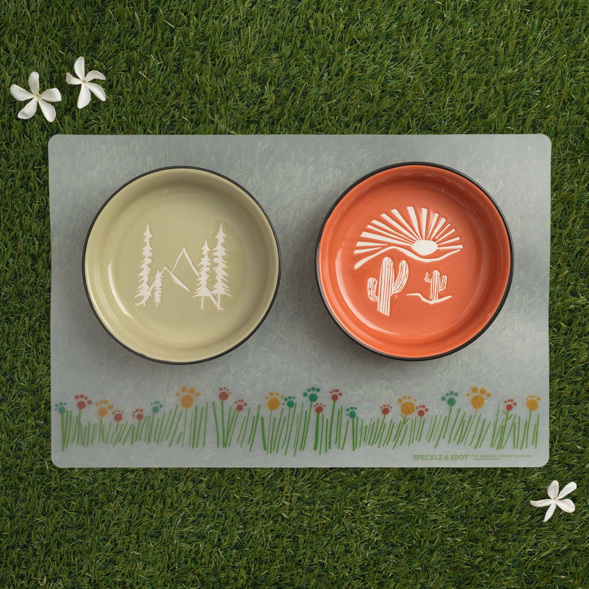 Two ceramic bowls with nature-themed designs on a placemat with floral border, placed on grass.
