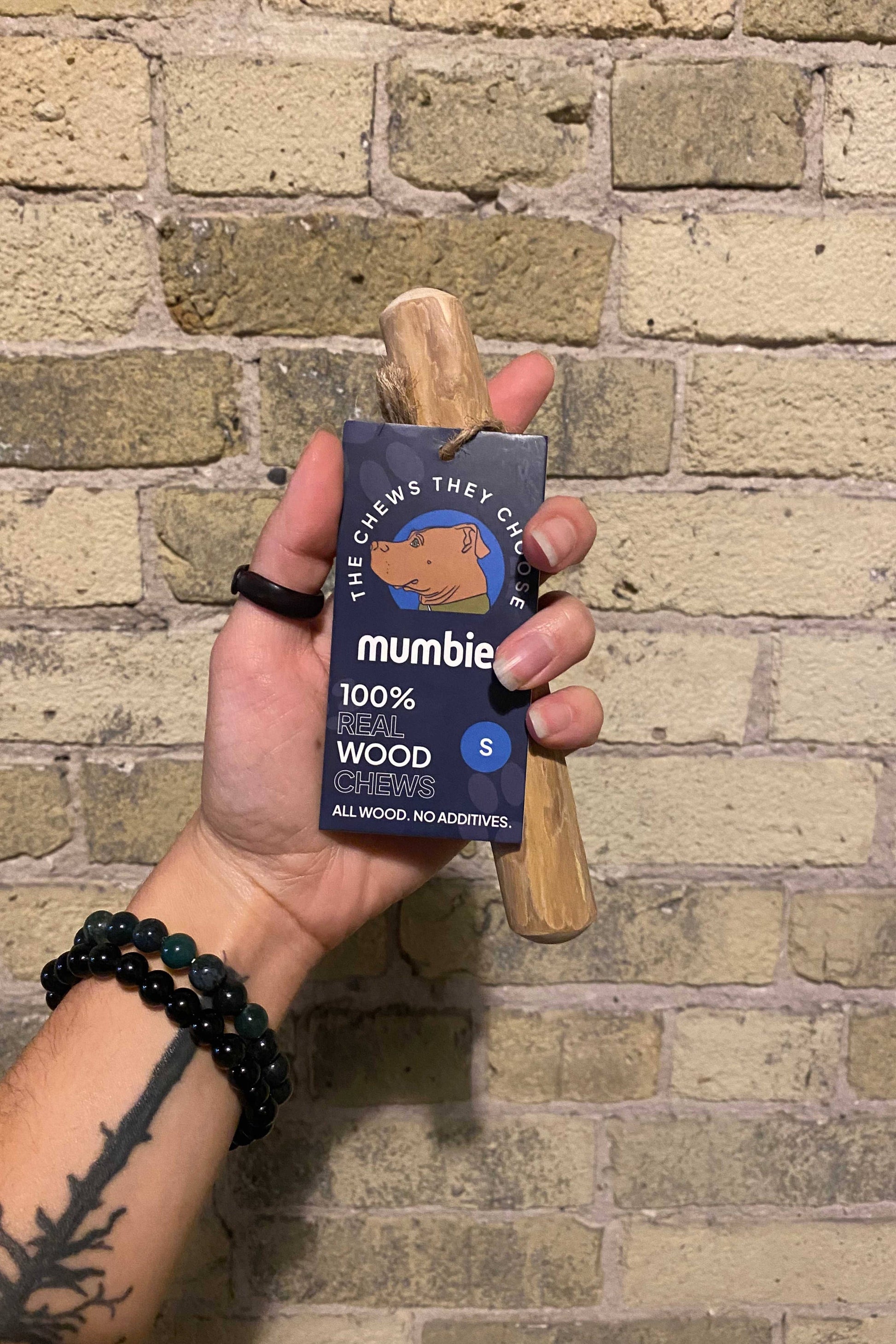 Hand holding a mumbie product with a wooden chew against a brick wall.