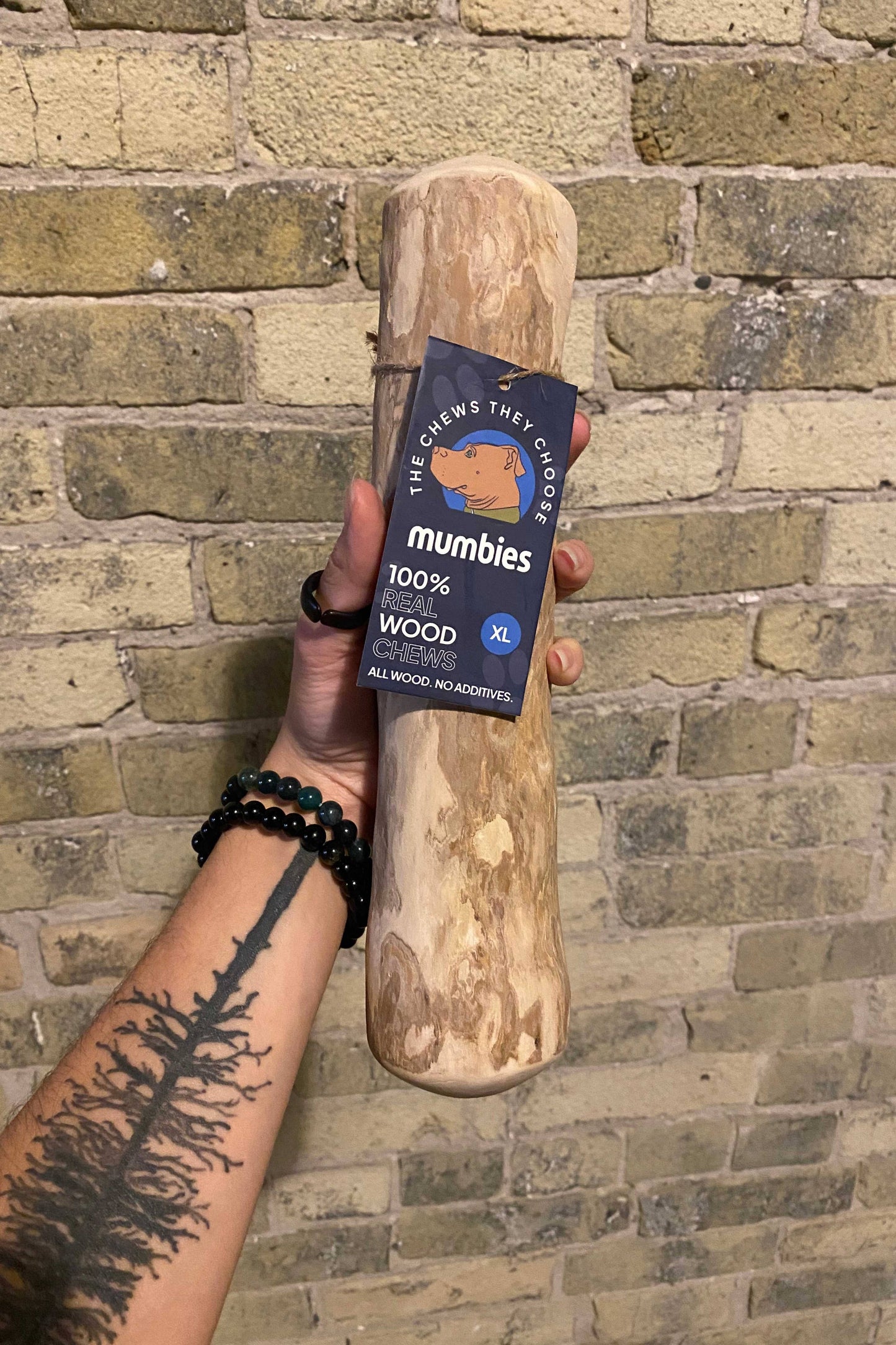 Hand holding a wooden chew toy with 'mumbies' branding against a brick wall.